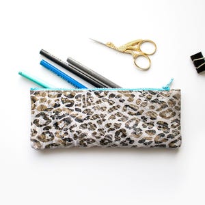 Op de afbeelding: Een rechthoekige etui met luipaardprint en een turquoise rits, met pennen en potloden. Een gouden schaar en een zwarte paperclip zijn ook zichtbaar.
