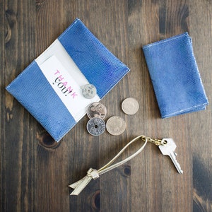 May include: A blue leather wallet with a white interior and a "Thank You!" card. Several coins and a key with a gold-colored keychain are next to the wallet. The items are on a dark wood surface.