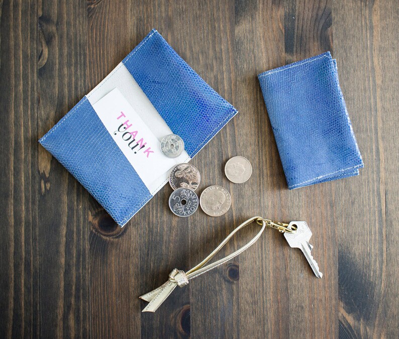 May include: A blue leather wallet with a white interior and a "Thank You!" card. Several coins and a key with a gold-colored keychain are next to the wallet. The items are on a dark wood surface.