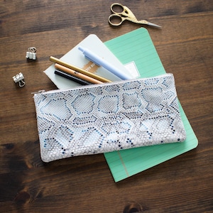 May include: A snakeskin-patterned pencil case in light blue and white, with a zip closure, on a green notebook. Gold scissors, pens, and binder clips are also visible on a dark wooden surface.