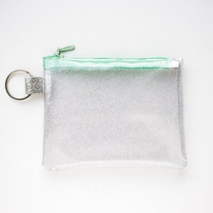 May include: A clear, rectangular glittery pouch with a mint green zipper and trim. The pouch has a silver key ring attached to the side. The pouch is designed to hold small items.