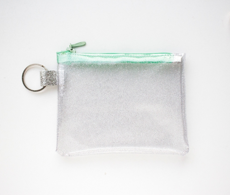 May include: A clear, rectangular glittery pouch with a mint green zipper and trim. The pouch has a silver key ring attached to the side. The pouch is designed to hold small items.