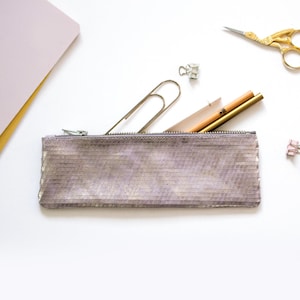 May include: A rectangular, zippered pencil case in shades of gray and gold, containing pencils, a paperclip, and a small notepad. Gold scissors and binder clips are scattered around the case on a white surface. Stationery supplies.
