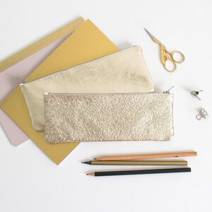 May include: Two gold-colored pencil cases with zippers, one with a textured surface. Also, a pair of gold scissors, pencils, and paper are visible. The items are arranged on a white surface, suggesting a stationery or office supply theme.
