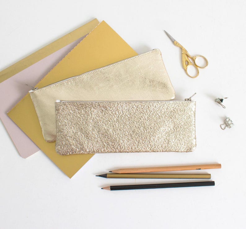 May include: Two gold-colored pencil cases with zippers, one with a textured surface. Also, a pair of gold scissors, pencils, and paper are visible. The items are arranged on a white surface, suggesting a stationery or office supply theme.