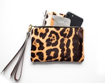 May include: A rectangular leopard print clutch with a wrist strap. The clutch is open, revealing a wallet and a black smartphone. The clutch has a gold zipper and a brown leather wrist strap.