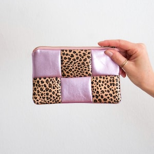 Könnte beinhalten: Eine rechteckige Reißverschlusstasche mit Patchwork-Design. Die Tasche hat abwechselnd Quadrate aus hellviolettem Metallic-Leder und Leopardenmuster-Stoff. Der Reißverschluss und die Nähte sind in einem passenden Hellrosa gehalten.