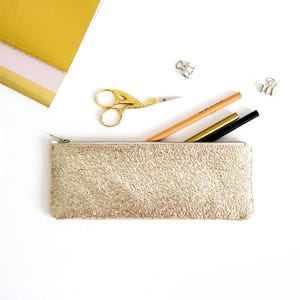 May include: A gold textured pencil case with a zipper, containing a pencil, a gold pen, and a black pen. A pair of gold scissors and binder clips are also visible. A gold notebook is in the background.