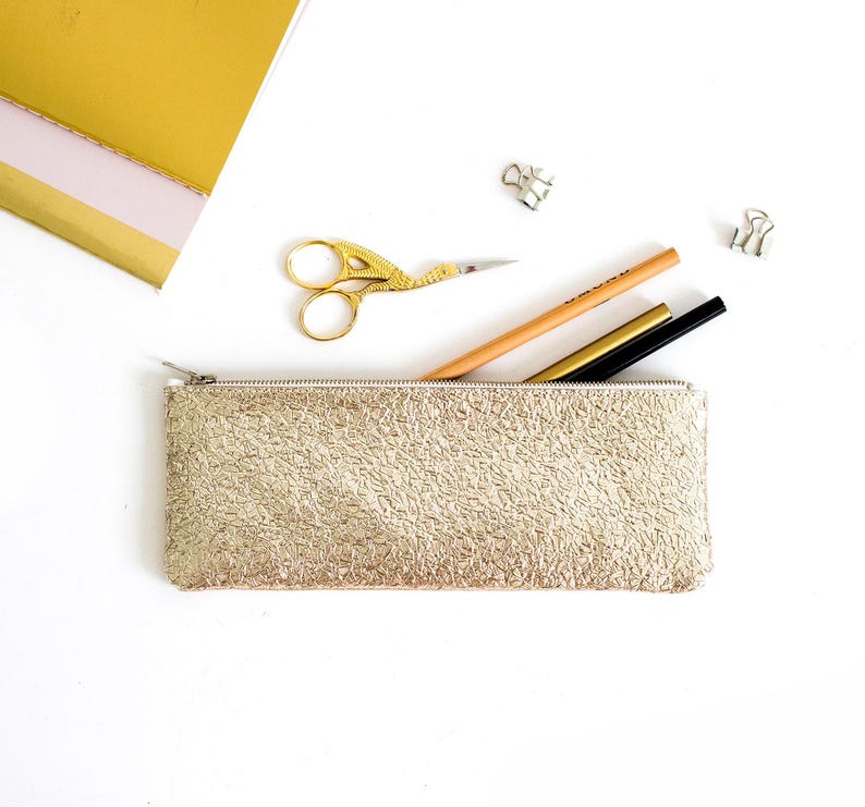May include: A gold textured pencil case with a zipper, containing a pencil, a gold pen, and a black pen. A pair of gold scissors and binder clips are also visible. A gold notebook is in the background.