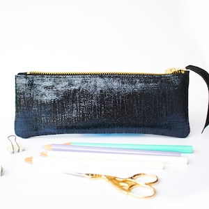 Puede incluir: Un estuche negro con cremallera dorada y una leng&uuml;eta de cuero negro. El estuche es rectangular y tiene una superficie texturizada. L&aacute;pices, tijeras y clips est&aacute;n colocados frente al estuche.