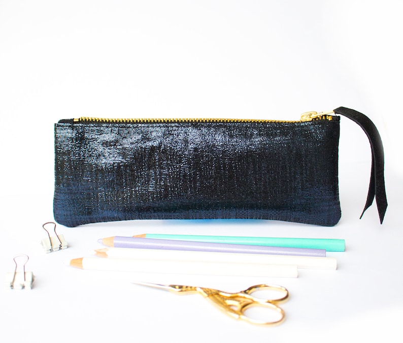 Puede incluir: Un estuche negro con cremallera dorada y una leng&uuml;eta de cuero negro. El estuche es rectangular y tiene una superficie texturizada. L&aacute;pices, tijeras y clips est&aacute;n colocados frente al estuche.