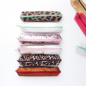 Può includere: Una collezione di astucci rettangolari in varie texture e colori, tra cui stampa leopardata, finta pelle di serpente bianca, rosa metallizzato, oro rosa, beige e bronzo, ciascuno con chiusura a zip sulla parte superiore.