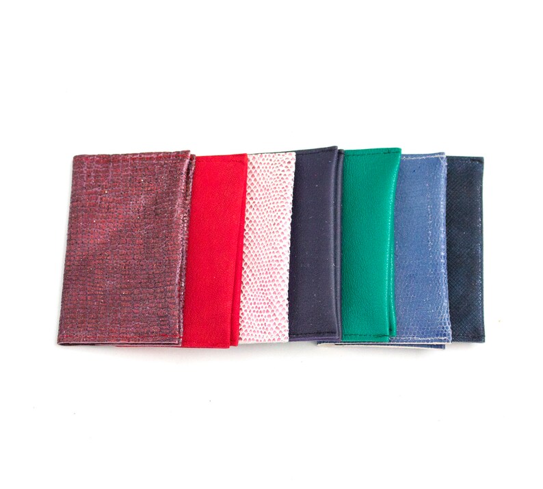 May include: A collection of colorful leather wallets in various shades, including burgundy, red, pink, navy, green, and blue. Each wallet has a unique texture or pattern, with a clean, minimalist design. The wallets are arranged in a row against a white background.