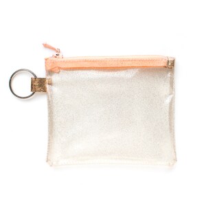 May include: A clear, glittery pouch with a peach-colored zipper and trim. The pouch has a silver key ring attached to a gold-colored fabric loop. The zipper pull is also peach-colored. The pouch is square-shaped.