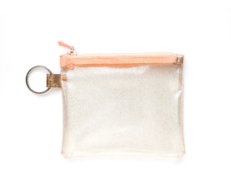 May include: A clear, glittery pouch with a peach-colored zipper and trim. The pouch has a silver key ring attached to a gold-colored fabric loop. The zipper pull is also peach-colored. The pouch is square-shaped.
