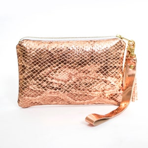 Puede incluir: Un bolso de mano en oro rosa con un estampado de piel de serpiente. El bolso rectangular tiene cierre de cremallera y una correa de muñeca con un cierre dorado y una pequeña borla. El bolso está hecho de un material brillante y texturizado.