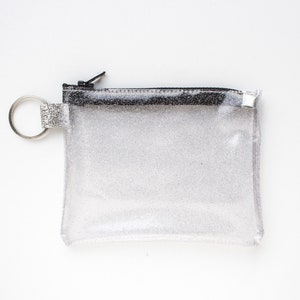 May include: A clear, glittery pouch with a black zipper and a silver key ring. The small, rectangular pouch is made of a transparent material with silver glitter throughout. The zipper is black, and the key ring is silver.