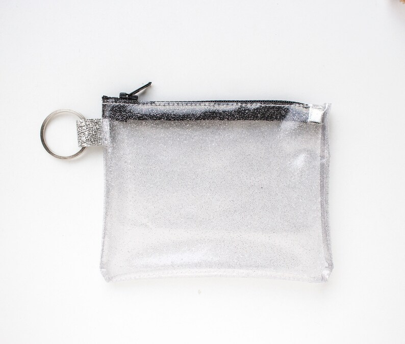 May include: A clear, glittery pouch with a black zipper and a silver key ring. The small, rectangular pouch is made of a transparent material with silver glitter throughout. The zipper is black, and the key ring is silver.