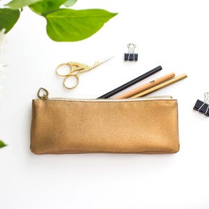 Op de afbeelding: Een goudkleurige leren etui met een rits, met potloden en een pen. Gouden scharen en paperclips liggen verspreid rond de etui op een wit oppervlak. Groene bladeren en witte bloemen zijn op de achtergrond.