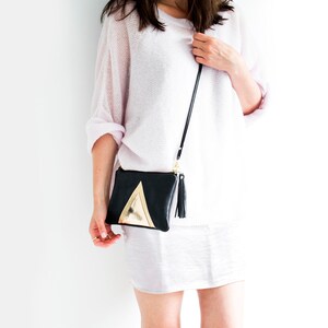 May include: Black crossbody bag with a gold triangle design and a black tassel. The bag has a long, thin black strap and a gold zipper. The bag is held by a person wearing a light pink sweater and skirt.