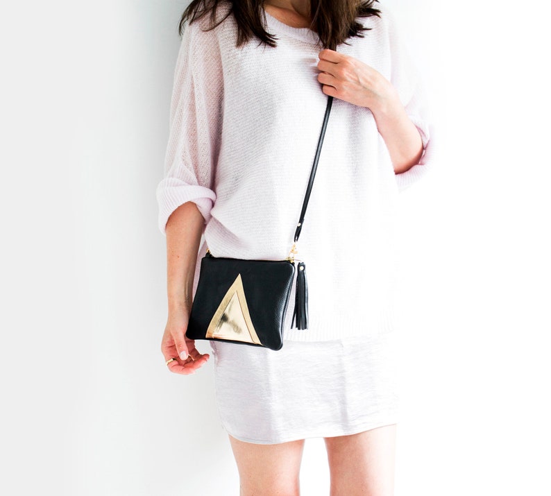 May include: Black crossbody bag with a gold triangle design and a black tassel. The bag has a long, thin black strap and a gold zipper. The bag is held by a person wearing a light pink sweater and skirt.