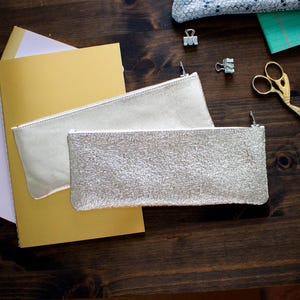 May include: Two silver pencil pouches with zippers are displayed on a dark wooden surface. One pouch has a smooth texture, while the other has a textured, crackled appearance. A gold notebook and other stationery items are also visible.