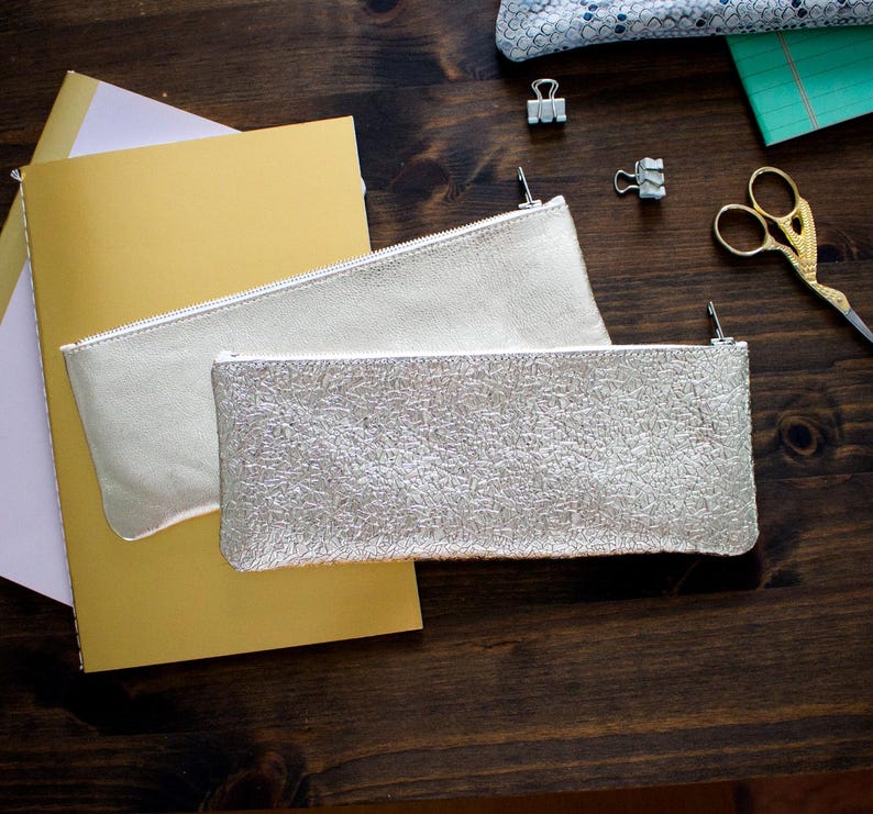 May include: Two silver pencil pouches with zippers are displayed on a dark wooden surface. One pouch has a smooth texture, while the other has a textured, crackled appearance. A gold notebook and other stationery items are also visible.