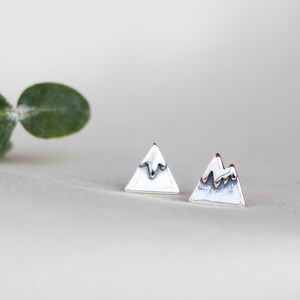 May include: A pair of silver mountain-shaped stud earrings. Each earring is a small, triangular shape with a textured, snowy peak. The earrings are set against a soft, neutral background, highlighting their minimalist design.
