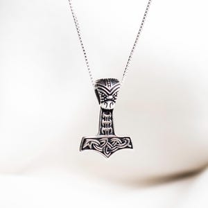 Könnte beinhalten: Eine silberfarbene Halskette mit einem detaillierten Hammeranhänger, der an einer feinen Kette hängt. Der Hammeranhänger weist komplizierte Muster und eine stilisierte Form auf, die wahrscheinlich von der nordischen Mythologie inspiriert ist. Die Halskette wird vor einem weichen, verschwommenen Hintergrund präsentiert.