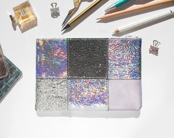 May include: A rectangular, patchwork-style pouch with a zipper closure. The pouch features sections of silver glitter, iridescent, and textured gray materials. Stationery items, including pencils and scissors, are scattered around the pouch on a white surface.