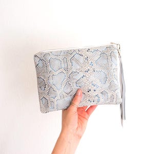 May include: A rectangular clutch with a snakeskin pattern in shades of light blue, gray, and white. The clutch has a wrist strap and a zipper closure. The bag is held against a white background.