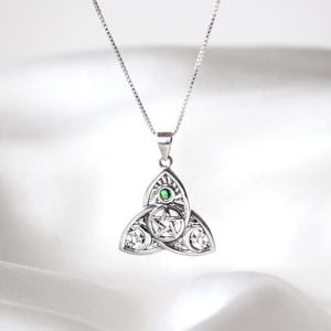 Op de afbeelding: Een zilverkleurige hangerketting met een Keltisch knoopontwerp en een pentagram in het midden. De hanger heeft een kleine groene edelsteen en hangt aan een fijne ketting. De achtergrond is een zachte, witte stof.