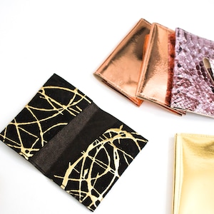 May include: A collection of stylish wallets in various colors and textures. The wallets include a black wallet with gold abstract designs, metallic copper, rose gold, and a textured pink snakeskin-like wallet. The wallets are displayed on a white background.