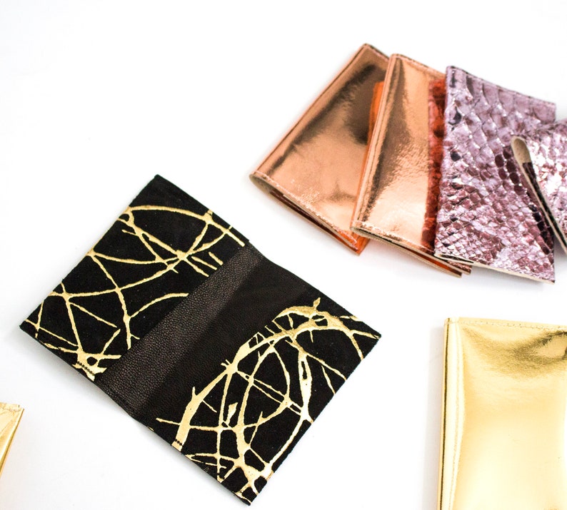 May include: A collection of stylish wallets in various colors and textures. The wallets include a black wallet with gold abstract designs, metallic copper, rose gold, and a textured pink snakeskin-like wallet. The wallets are displayed on a white background.