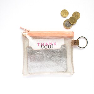 May include: A clear, rectangular pouch with a peach-colored zipper and a silver card holder. The pouch contains a card that says "THANK YOU!" in pink letters. A few euro coins are scattered nearby.