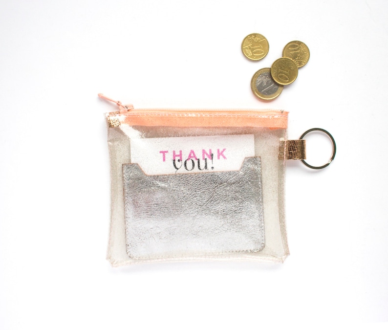May include: A clear, rectangular pouch with a peach-colored zipper and a silver card holder. The pouch contains a card that says "THANK YOU!" in pink letters. A few euro coins are scattered nearby.