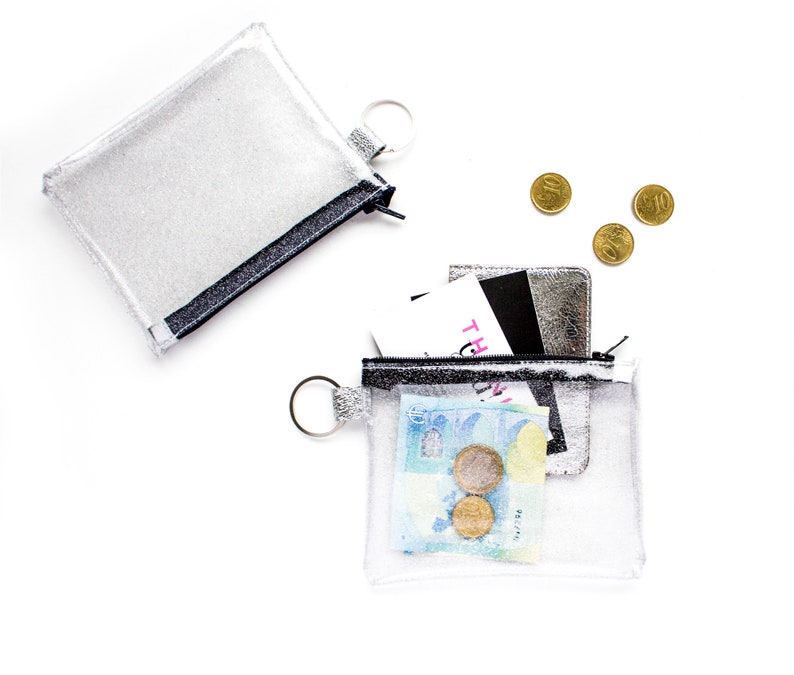 May include: Two clear, sparkly pouches with black trim and silver key rings. One pouch is closed, the other is open, revealing money, cards, and coins. The background is white.