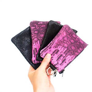 May include: A hand holds a fan of four small, rectangular coin purses. Two are black with a textured pattern, and two are pink with a similar pattern. Each purse has a zipper and a silver key ring.