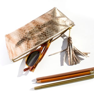 Könnte beinhalten: Eine gold- und beigefarbene Reißverschlusstasche mit Schlangenmuster und passendem Quastenanhänger, in der sich eine Sonnenbrille mit Schildpattmuster befindet. Drei Bleistifte in Braun- und Goldtönen sind darunter angeordnet.