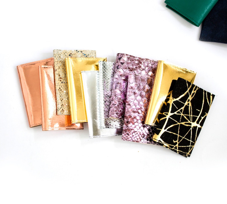 May include: A collection of passport holders in various colors and textures. The holders include metallic gold, silver, and rose gold, along with a black holder with gold abstract lines and a pink snakeskin pattern. The holders are arranged on a white surface.