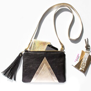 May include: Black and gold crossbody bag with a geometric design. The bag has a long strap, a black tassel, and a zipper closure. A phone and a small, sparkly wallet are inside. A keychain with a "Thank You!" tag and keys are also present.