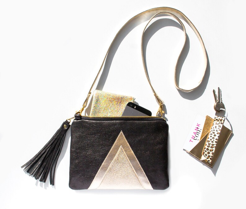 May include: Black and gold crossbody bag with a geometric design. The bag has a long strap, a black tassel, and a zipper closure. A phone and a small, sparkly wallet are inside. A keychain with a "Thank You!" tag and keys are also present.