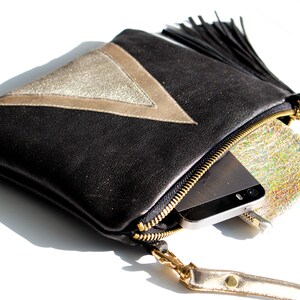 May include: Black leather clutch with a gold metallic triangle design and a black tassel. The clutch has a gold zipper and a gold wrist strap. A smartphone and a small silver pouch are inside.