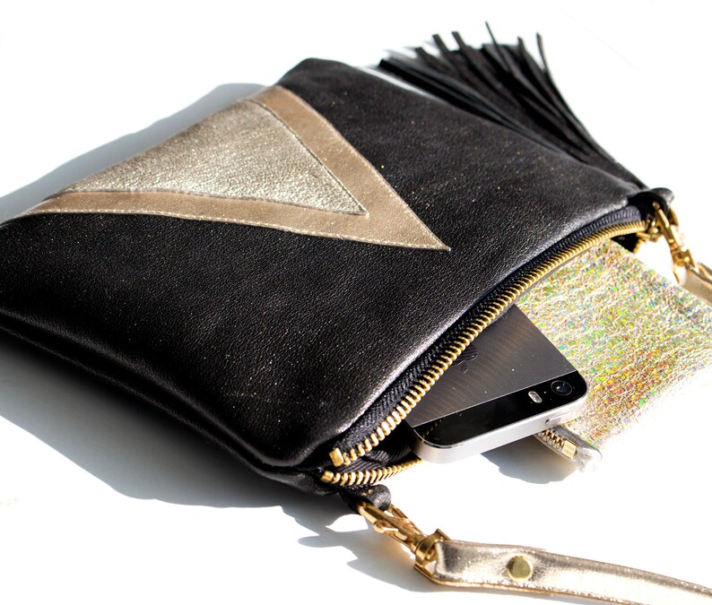 May include: Black leather clutch with a gold metallic triangle design and a black tassel. The clutch has a gold zipper and a gold wrist strap. A smartphone and a small silver pouch are inside.