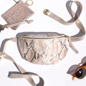 Może przedstawiać: Zestaw akcesoriów, w tym nerka ze wzorem skóry węża ze złotym zamkiem błyskawicznym, mały brokatowy różowy etui na karty z kluczami i okulary przeciwsłoneczne w kolorze żółwiej skorupy. Nerka ma złoty pasek.