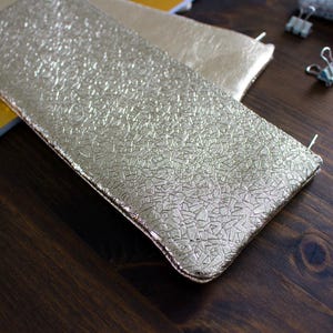 May include: Gold-colored rectangular pouch with a textured surface. The pouch is made of a shiny material and has a zipper closure. Another similar pouch is visible in the background.