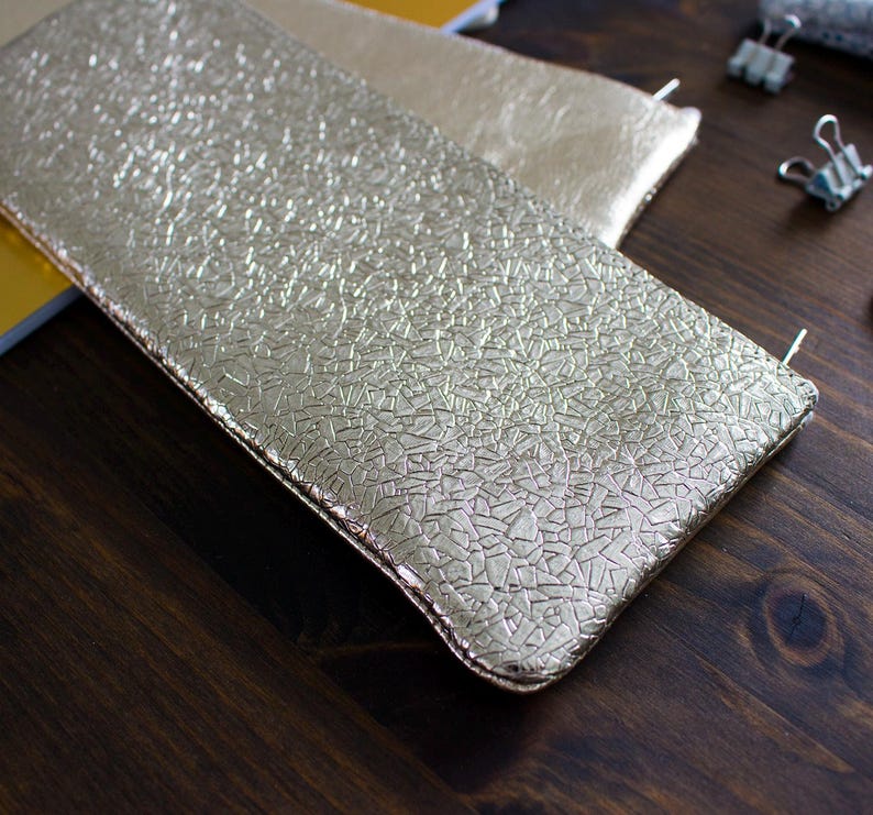 May include: Gold-colored rectangular pouch with a textured surface. The pouch is made of a shiny material and has a zipper closure. Another similar pouch is visible in the background.