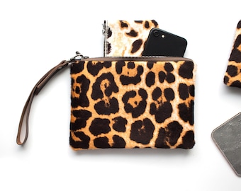 May include: A leopard print clutch with a brown wrist strap and a black smartphone inside. The clutch is made of a textured material with a zipper closure. Additional leopard print items are visible.