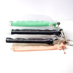May include: Four small, clear zippered pouches with silver key rings. The pouches have different colored zippers: mint green, black, and peach. The pouches are stacked on top of each other against a white background.
