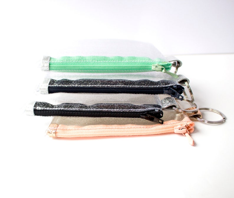 May include: Four small, clear zippered pouches with silver key rings. The pouches have different colored zippers: mint green, black, and peach. The pouches are stacked on top of each other against a white background.